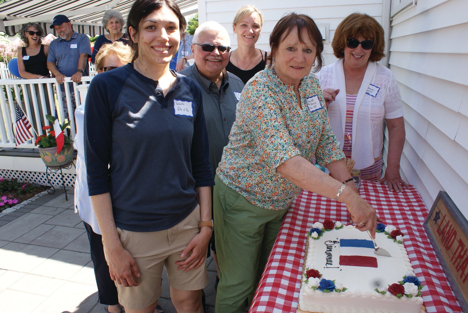 IT’S A PICNIC: Supportive friends, neighbors and family happily join together to greet their guests from France. At right Pierrete cuts into the ceremonial welcome cake decorated with the French flag as Laura, Ed, Mylene and family Catherine Conant look on.